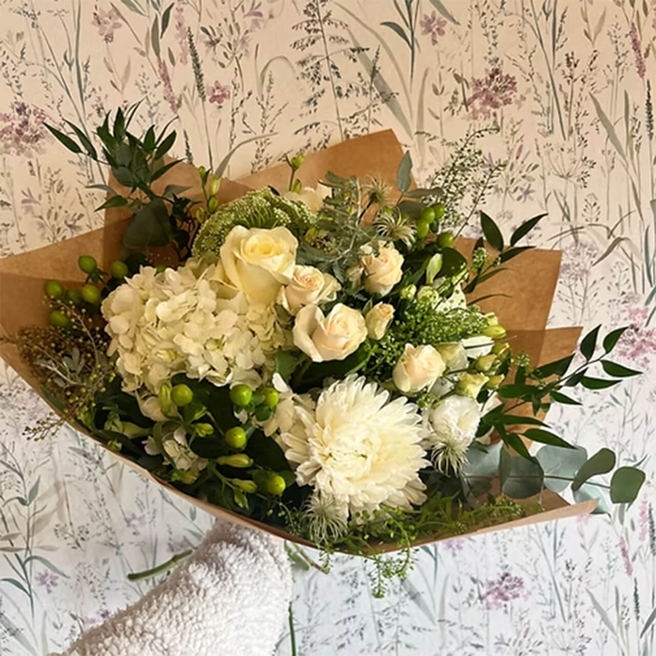 Hand-tied bouquet of white roses, hydrangea, chrysanthemum and green hypericum berries wrapped in brown kraft paper