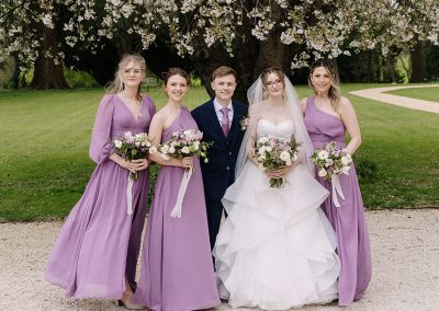 Bride with two bridesmaids in lilac dresses holding matching bouquets of white and purple flowers, outside a country house wedding venue