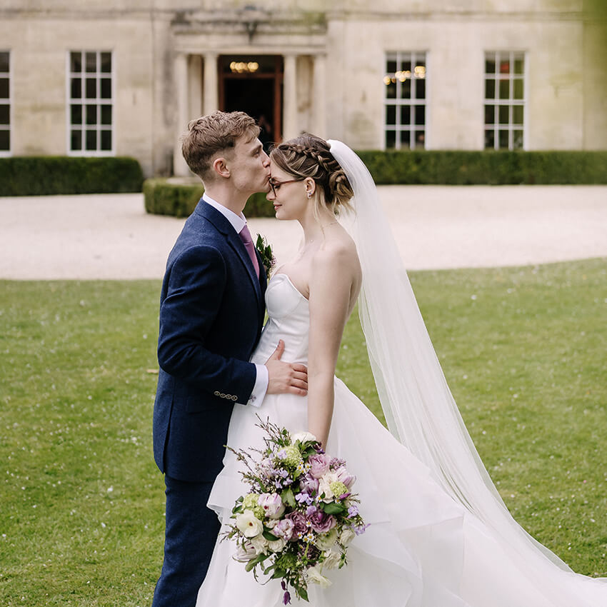 Bride and groom sharing a kiss outside a country house wedding venue, bride holding a trailing bouquet of white and pink flowers by The Flower Shed Westerleigh