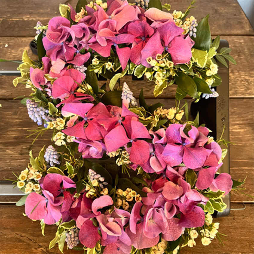 Fresh floral letter S arrangement featuring vibrant pink hydrangeas, pale yellow waxflowers, light blue muscari grape hyacinths, and variegated green foliage