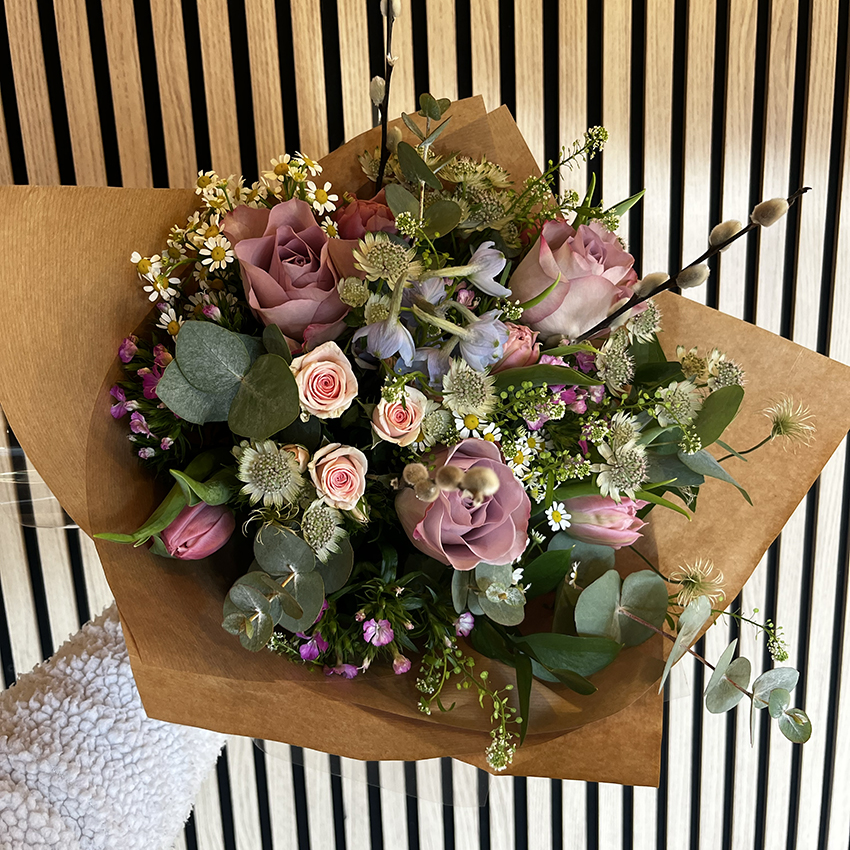 Hand-tied floral bouquet wrapped in brown kraft paper featuring dusty pink roses, miniature pink rosebuds, chamomile daisies, astrantia, and eucalyptus sprigs