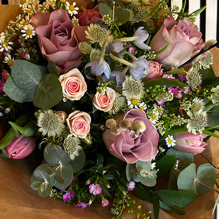 Hand-tied floral sheath for funeral wrapped in brown kraft paper featuring dusty pink roses, miniature pink rosebuds, chamomile daisies, astrantia, and eucalyptus sprigs