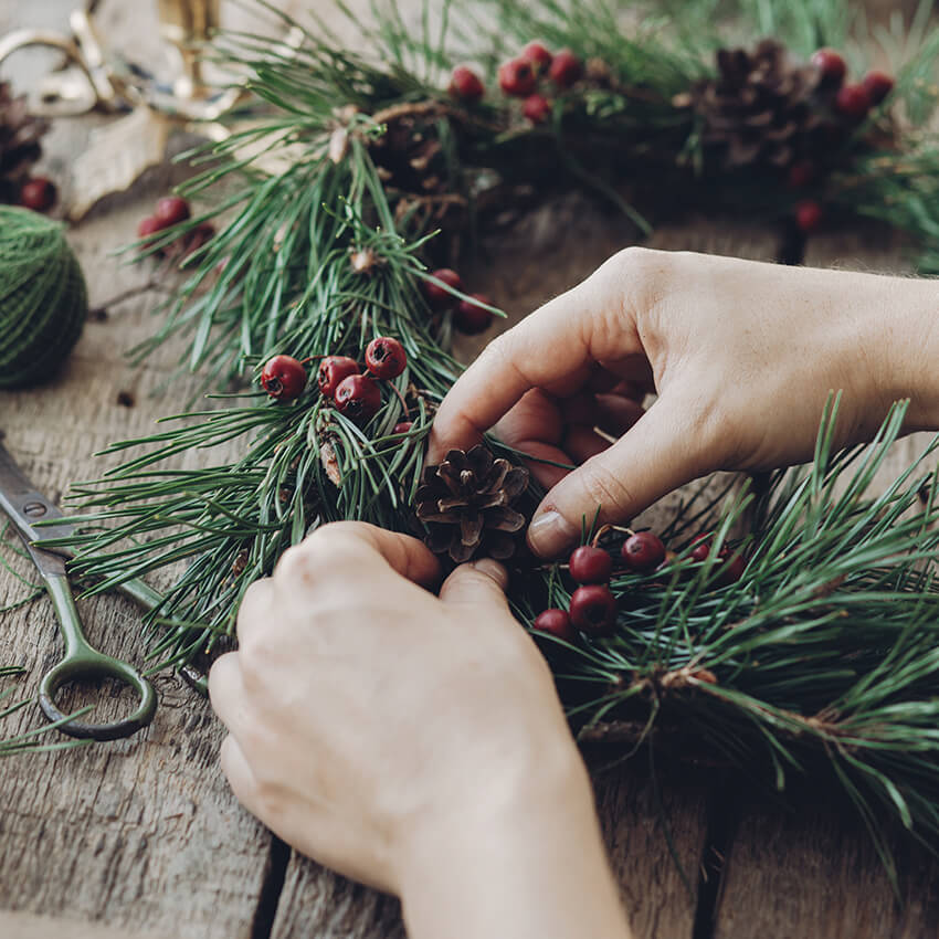 Overhead view of hands assembling a DIY Christmas wreath with fresh green pine needles, small pinecones, and red winter berries on a rustic wooden table.