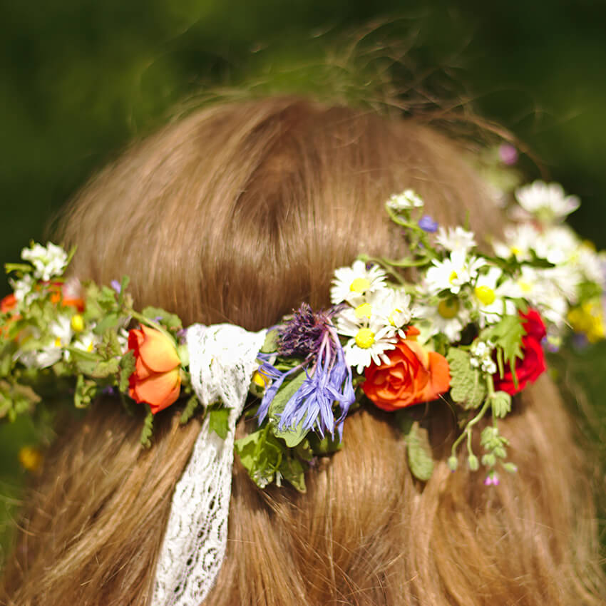 Rustic boho bridal flower crown featuring fresh wildflowers, vibrant orange spray roses, and a hanging white lace tie for a summer wedding style.