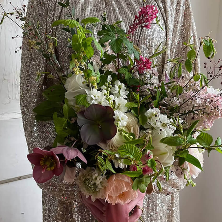 A close-up view of a bride or bridesmaid in a sparkling gold sequin dress holding an organic, hand-tied spring bridal bouquet. The textured arrangement features dark purple and pink hellebores, white delphiniums, peach-toned poppies, bright pink flowering currant stems, and fresh green raspberry foliage, finished with a dusty rose silk ribbon wrap.