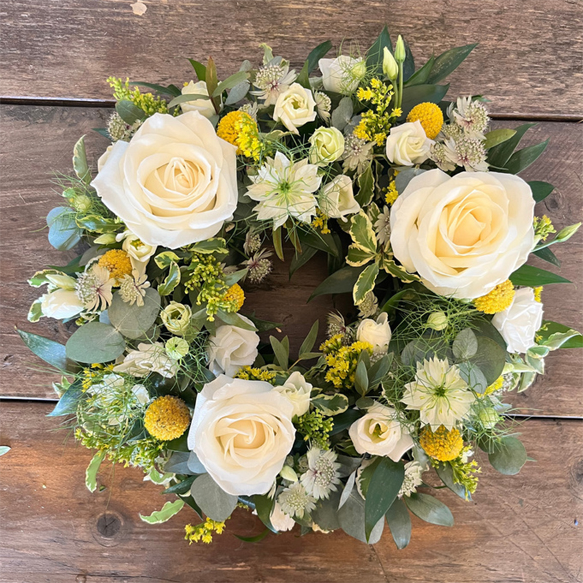 A circular funeral wreath tribute laid flat on a rustic wooden background. This sympathy flower arrangement features large white roses symbolising remembrance, vibrant yellow craspedia billy buttons, star-shaped white nigella flowers, and pale green astrantia, all woven with comforting green eucalyptus foliage.
